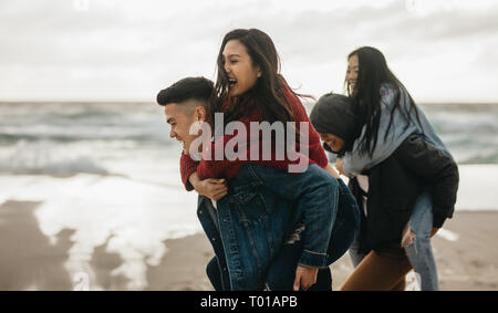 Giovani uomini che trasportano gli amici femmina sul loro dorso lungo il mare. Gruppo di amici piggybacking all'aperto sulla riva del mare. Foto Stock