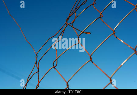 Rotto e danneggiato opaco filo di maglia recinto con un cielo blu e un intero nella scherma Foto Stock
