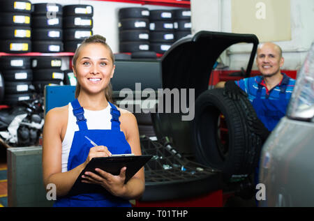 Giovane femmina felice tecnico nella tuta negli appunti di contenimento in officina per auto Foto Stock