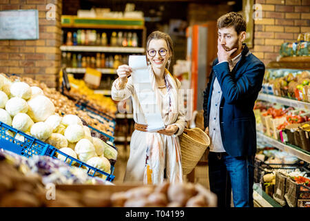 Coppia giovane comprare generi alimentari in piedi insieme con una lunga lista di shopping nel reparto vegetali del supermercato. Uomo con scioccato emozioni Foto Stock
