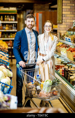 Ritratto di una giovane e felice coppia in piedi con carrello pieno di cibo al supermercato Foto Stock