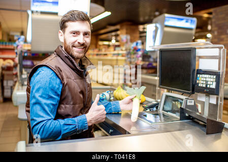 Ritratto di un felice e allegro uomo come un cassiere, seduto alla cassa al supermercato Foto Stock
