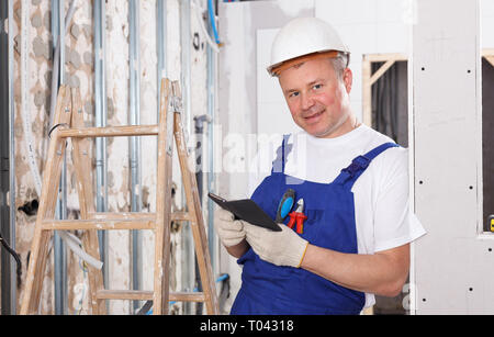 Contraente maschio avente pausa a interni cantiere, appoggiata alla parete con il telefono in mano Foto Stock