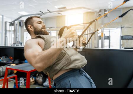 Muscolare uomo barbuto vestito di ponderata militare giubbotto corazzato facendo esercizi utilizzando sistemi di cinghie in palestra. Sport, formazione bodybuilding e guarire Foto Stock
