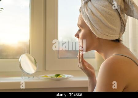 Giovane donna in casa vicino alla finestra guardando in uno specchio, la cura per il viso con crema e facendo una maschera con il cetriolo e limone, asciugamano sulla testa. Cura della pelle, c Foto Stock