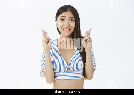 Pregando per il sogno. Speranzoso ambizioso bella donna con capelli scuri in alto ritagliata incrocio le dita per la buona fortuna di mordere il labbro inferiore mentre Foto Stock