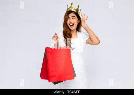 Stupiti donna asiatica in Golden crown holding mazzetto di sacchetti di carta mentre in piedi su sfondo bianco Foto Stock