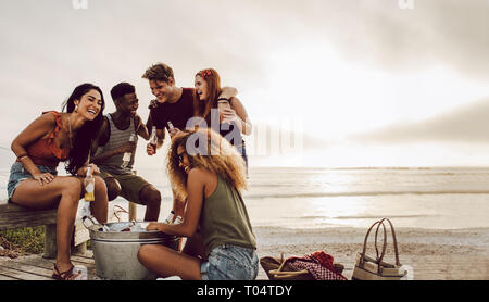 Gruppo di multi-etnico amici gustando birre insieme alla spiaggia. Un gruppo di uomini e donne a bere birra in riva al mare. Foto Stock