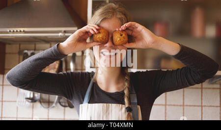 Ritratto di bella donna in grembiule che copre la sua gli occhi con muffin e sorridere mentre in piedi in cucina Foto Stock