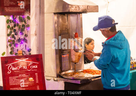 Uomo che indossa abiti blu e cappuccio preparazione shwarma su una griglia verticale in corrispondenza di un cibo all'aperto chiosco Foto Stock