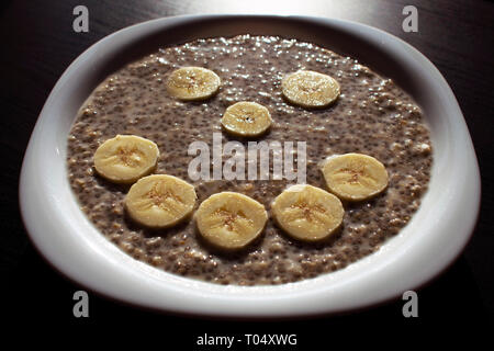 Semi di Chia e fiocchi di avena in acqua, con fettine di banana disposte in faccina sorridente su una piastra bianca. Colazione perfetta. Foto Stock