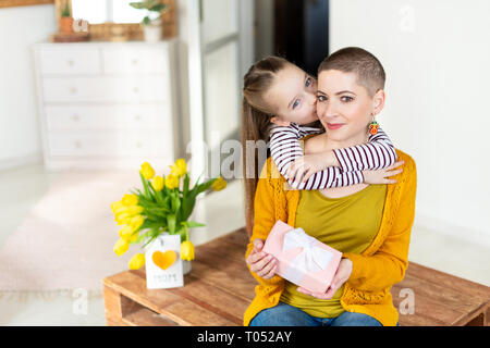 Felice Festa della mamma Festa di compleanno o di sfondo. Adorabile ragazza giovane sorprende la sua mamma, giovane malato di cancro, con bouquet con il presente. Festa di famiglia Foto Stock