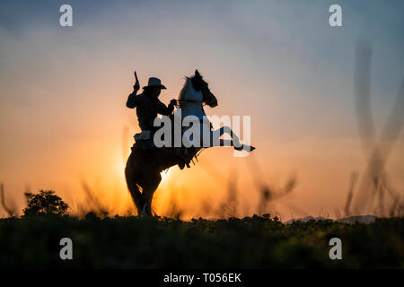 Silhouette cowboy sparare una pistola su un cavallo in sunset Foto Stock