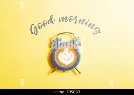 Orologio sveglia con il quadrante di cappuccino su sfondo giallo. In stile minimale di caffè il concetto di tempo con iscrizione Buongiorno. Foto Stock
