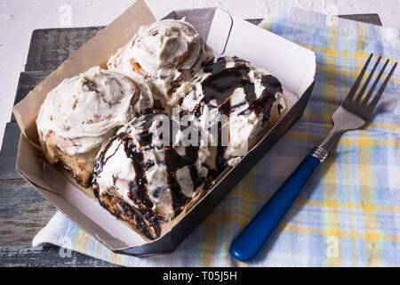 Piatto di laici Cinnabons - panini alla cannella con crema di formaggio e di cioccolato, in una scatola di carta, oltre il tovagliolo e legno sfondo blu. Foto Stock