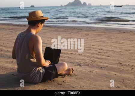 Vista posteriore dei piedi nudi uomo in hat seduto sulla spiaggia di sabbia vicino a sventolare il mare e la navigazione portatile moderno Foto Stock