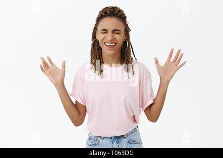 Divertito e gioioso di spensierata donna afro-americana con i timori in rosa t-shirt chiudendo gli occhi dei bracci di sollevamento con palme facce a camear sorridente con chiuso Foto Stock