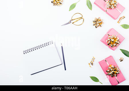 Festa laica piatta disposizione del blocco note vuote nei pressi di rosa confezioni regalo e decorazioni dorate sul tavolo bianco, vista dall'alto. Foto Stock