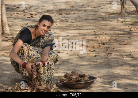 Donna rurale vestito in saree raccolta di foglie secche dal terreno in un ferro da stiro coppa d'oro. Foto Stock