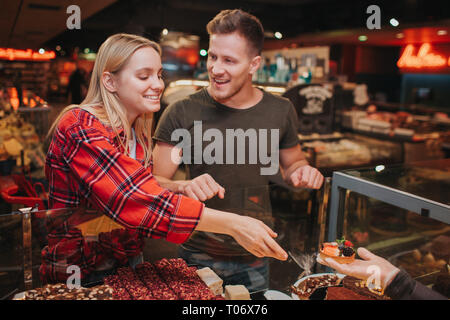 Coppia giovane in un negozio di alimentari. Piacevole giovane donna a ottenere la torta dal lato e sorriso. Ella ha portata di mano. Giovane uomo guarda il suo sorriso e. Foto Stock