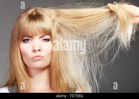 Cura dei capelli -. Donna bionda con la sua danneggiato i capelli secchi volto arrabbiato espressione sfondo grigio Foto Stock