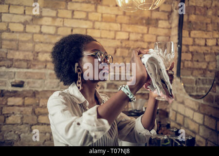 Bella-attraente dai capelli scuri afro-americani interessati cameriera facendo vino lucidatura di vetro Foto Stock