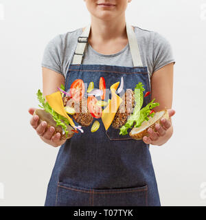 Lo chef femmina in un grembiule in denim con battenti gustosi hamburger fatti in casa da freschi ingredienti naturali nelle sue mani su uno sfondo bianco. Posto per il testo. Foto Stock