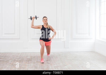 Ritratto di giovane atletico culturista bella donna in pantaloncini rosa e nero in alto facendo squat e gli esercizi in palestra con i manubri su bianco Foto Stock