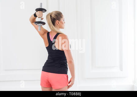 Sforzandosi per forte. Retro del giovane adulto motivazione athletic culturista donna in pantaloncini rosa e nero in alto standing, holding manubri facendo exerc Foto Stock