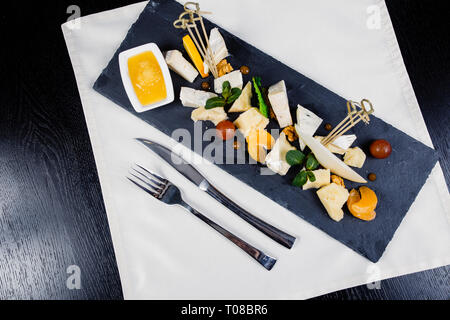 Il piatto di formaggi assortimento di vari tipi di formaggio su ardesia scura scheda di taglio. Formaggio Camembert, rococò, parmigiano, dadi, pomodori ciliegini, mandarino, o Foto Stock