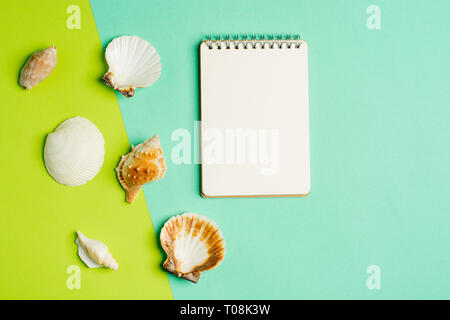 Vacanze estive piana concetto laici con il blocco note di bianco e conchiglie sulla trendy pastello blu-verde dello sfondo. Vista dall'alto. Foto Stock