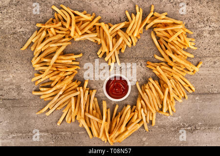 Chiudere fino a forma di cuore di patate fritte isolato su sfondo concreto, ketchup ciotola nel mezzo. Direttamente al di sopra. Foto Stock