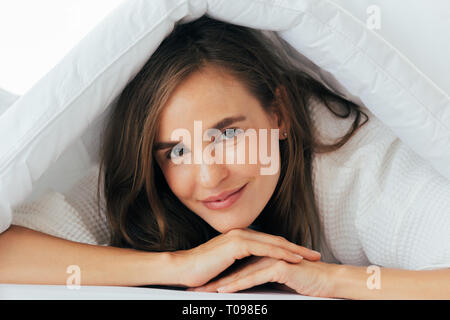 Giovani calma e tranquilla donna caucasici in bianco pigiami guardando la fotocamera e la copertura lei stessa con coperta in camera da letto Foto Stock