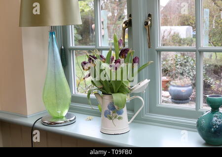 Un davanzale di legno con lampada in vetro, una lattina di metallo con tulipani sbiadito e un vaso decorativo. Un giardino può essere intravisto attraverso la finestra in legno. Foto Stock