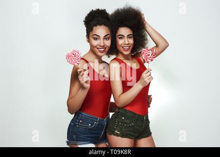 Dolce bellezza. Due belle giovani afro american donne in abiti estivi di contenimento a forma di Lecca Lecca Cuore e sorridere mentre in piedi contro backgro grigio Foto Stock