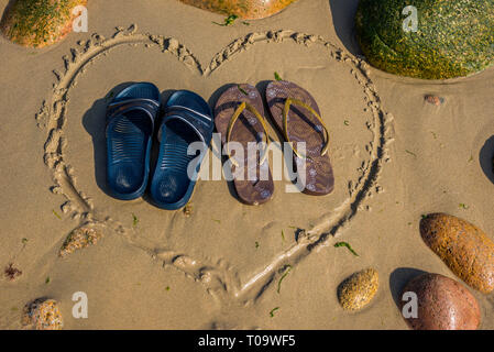 Una coppia di elementi maschio e un paio di calzature femminili in piedi a forma di cuore sulla spiaggia Foto Stock