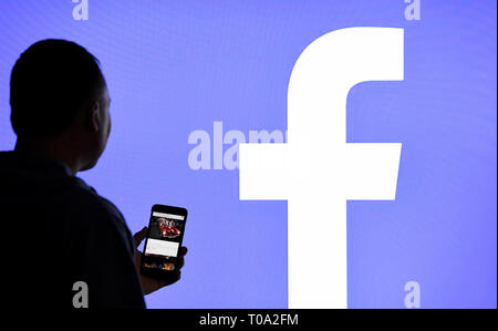 15 marzo 2019, Schleswig-Holstein, Aukrug-Homfeld: Illustrazione - un uomo con uno smartphone sta in piedi di fronte ad un monitor con il logo di Facebook. Foto: Carsten Rehder/dpa Foto Stock