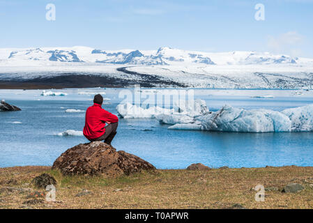 L'uomo traveler in giacca rossa si trova sulla riva di Jokulsarlon laguna glaciale. Paesaggio estivo, di ghiacciai e iceberg di Islanda. Parco nazionale di Vatnajoku Foto Stock