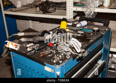 Scatola con strumenti e parti di ricambio giacente su di esso in un disordine nel processo di riparare la vettura in officina per la manutenzione del veicolo Foto Stock