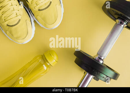 Piatto di laici una bottiglia di acqua minerale, sneakers e manubri su sfondo giallo fitness minimo concetto creativo. Foto Stock