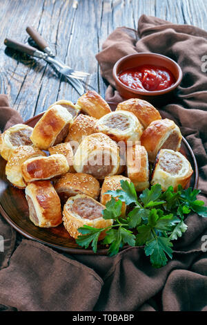 Salsiccia rotola su una piastra di terracotta su legno scuro tavolo rustico con panno marrone e salsa di pomodoro in una ciotola, finger food, cucina inglese, vertica Foto Stock