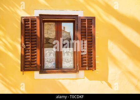 Una splendida finestra con persiane di legno in giallo sulla parete luminosa giornata estiva con albero ombra sul muro Foto Stock