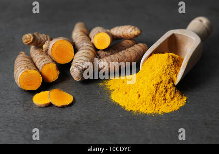 La curcuma in polvere di spezie sano cibo asiatico closeup di curcuma radici affettato e un depositante di legno su un rustico grigio scuro scheda di cucina. Foto Stock