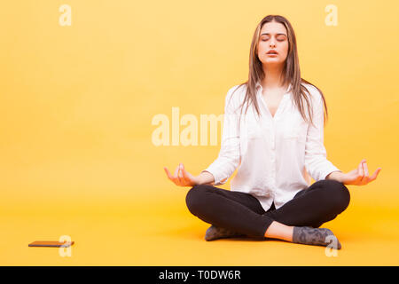 Ragazza millenario di prendere una pausa dal suo telefono e la pratica di meditazione su sfondo giallo. Stato pacifico della mente Foto Stock