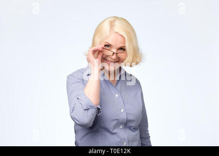 Senior donna occhiali di regolazione e sorridente a voi in piedi su sfondo bianco Foto Stock