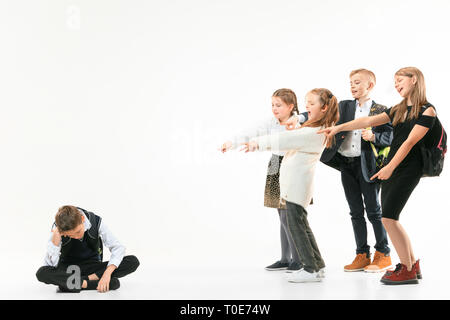 Piccolo Ragazzo seduto da solo sul pavimento e la sofferenza di un atto di bullismo mentre i bambini beffardo in background. Triste giovane scolaro seduta su studio contro uno sfondo bianco. Foto Stock