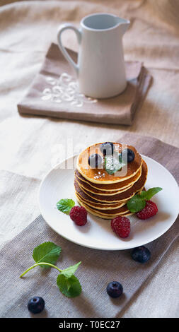 Frittelle con frutti di bosco i frutti di bosco sul tavolo bianco con il bricco per il latte. I pancake freschi sulla piastra tovaglia. I mirtilli, lamponi, menta sulle frittelle. Foto Stock