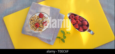Sana colazione con vetro muesli fatti in casa con yogurt e frutti di bosco freschi lamponi in argento tazza succo d'arancia fresco menta grigio panno da cucina tovagliolo su sfondo giallo. Messa a fuoco selettiva, vista dall'alto Foto Stock