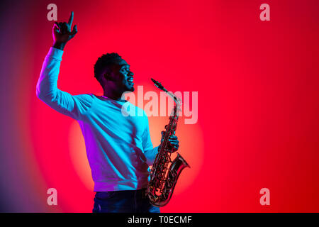 African American bello il musicista jazz interagisce con il pubblico e tenendo il sassofono in studio su un sfondo al neon. Il concetto di musica. Giovani gioiosi ragazzo attraente improvvisato. Close-up ritratto retrò. Foto Stock