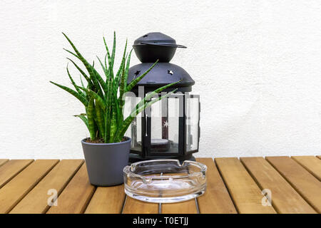 Vecchia lanterna in vetro con candela, Sansevieria cylindrica (cilindrico di serpente) Vegetali di fiori in vaso e posacenere sul tavolo in legno da muro bianco sullo sfondo. Ho Foto Stock